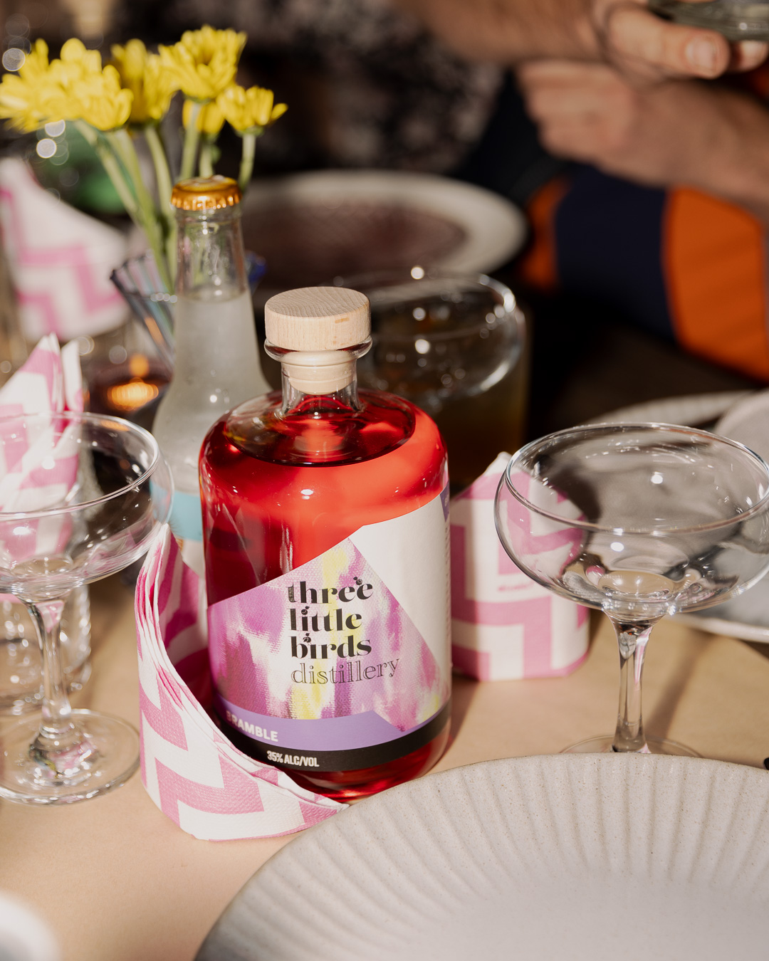 Bottle of Three Little Birds Bramble Gin on a festive table with pink napkins, coupe glasses and yellow flowers