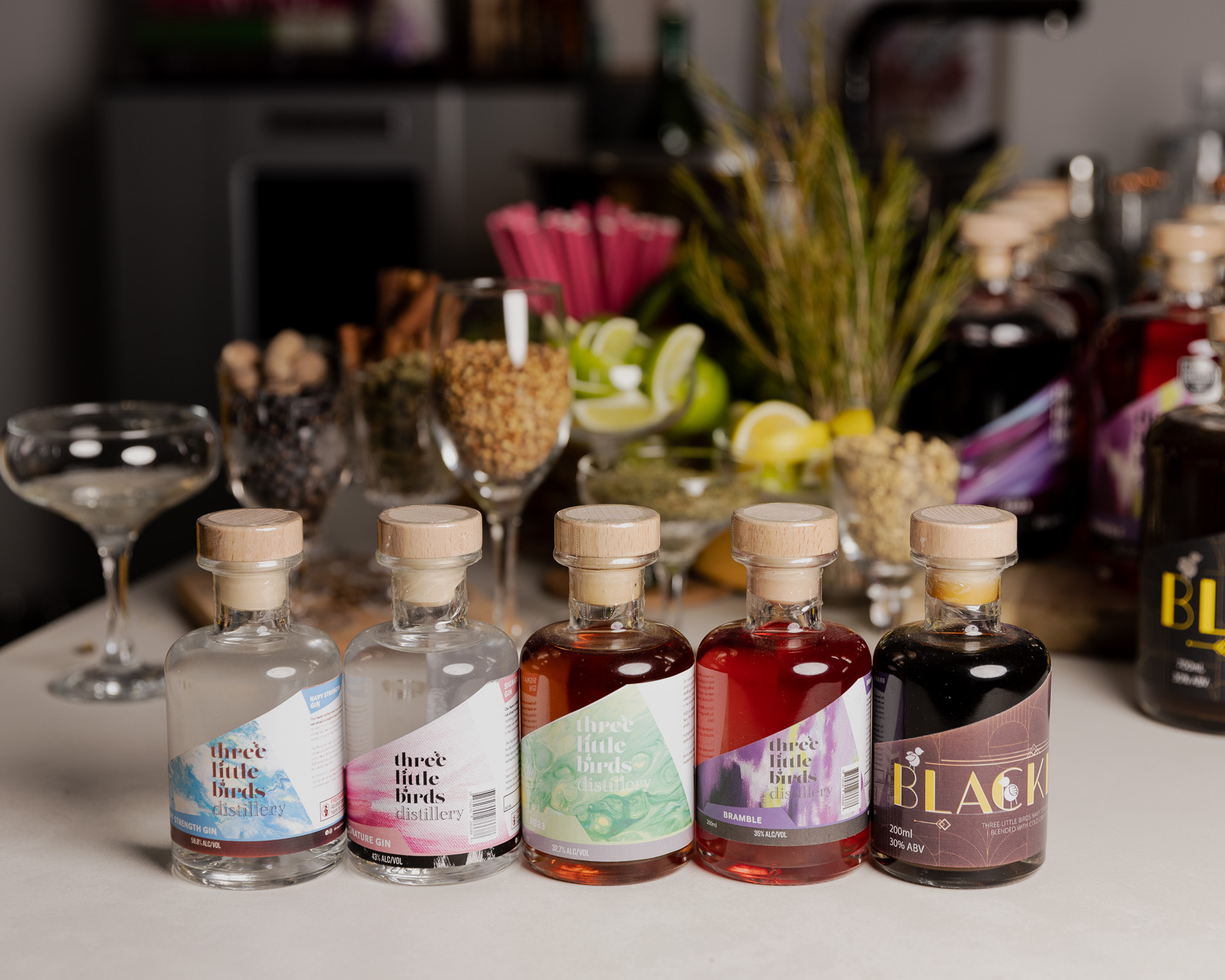 A lineup of Three Little Birds gin bottles on the tasting room bar, botanicals and citrus behind them.